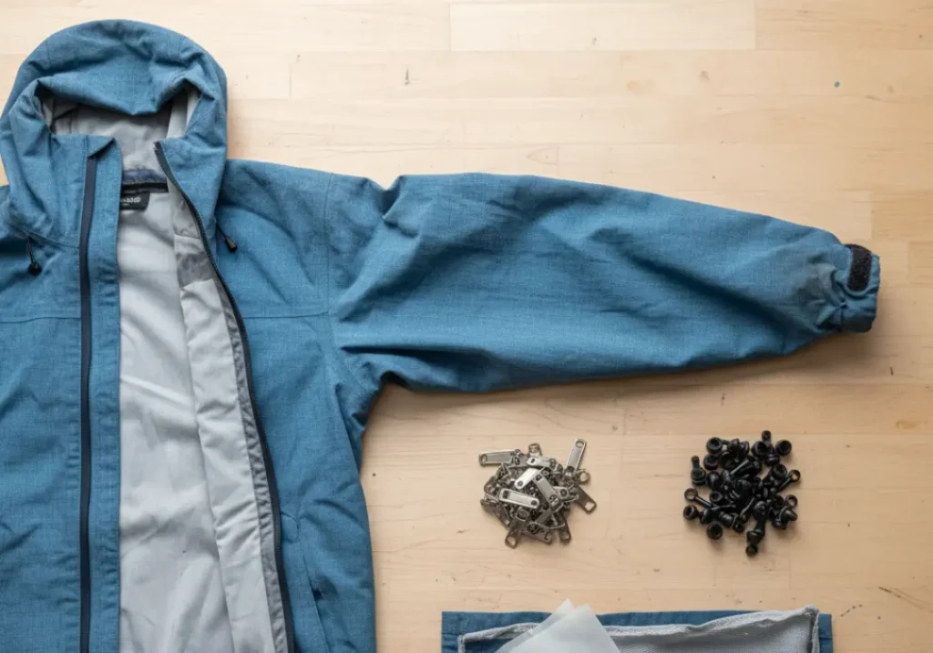 A deconstructed hiking jacket on a table, with its zippers, buckles, and fabric layers sorted into separate piles for recycling.