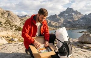 The 2026 Master Guide to Thru Hike Resupply Box Logistics A pristine thru-hiker sitting on a granite peak in the Sierra Nevada, organizing a resupply box filled with high-end hiking food, wearing a red Arc'teryx jacket and next to a white Hyperlite Mountain Gear pack, bathed in golden hour sunlight.