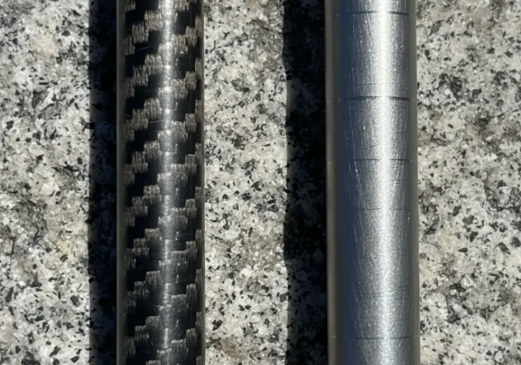 A close-up comparison of a carbon fiber trekking pole's woven texture next to the matte finish of an aluminum trekking pole on a rock.