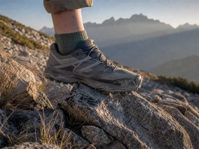 0-rock-plate-hiking-shoes-rocky-trail Low-cut hiking shoe stepping onto sharp granite on a rocky trail at first light, sole flexing over a pointed rock