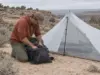 Silnylon vs Dyneema (DCF) What Fails First on Trail Hiker packing gear beside a pitched DCF shelter on a sandy desert bench, silnylon vs Dyneema choice in the field