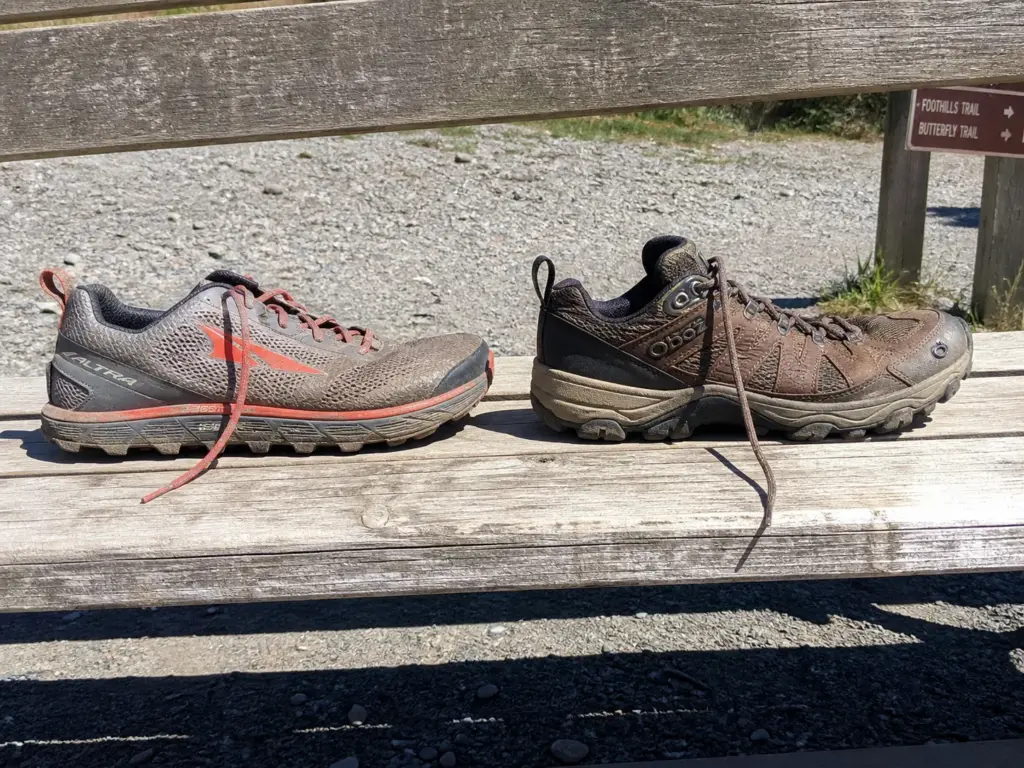 Side view comparing a zero-drop trail shoe and a traditional hiking shoe on a wooden bench