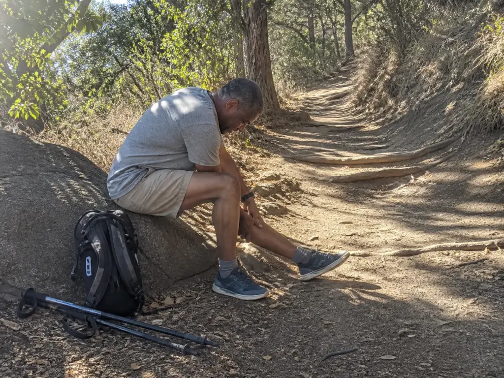 Hiker seated on a trail rock reaching toward his calf after a hike in zero-drop shoes