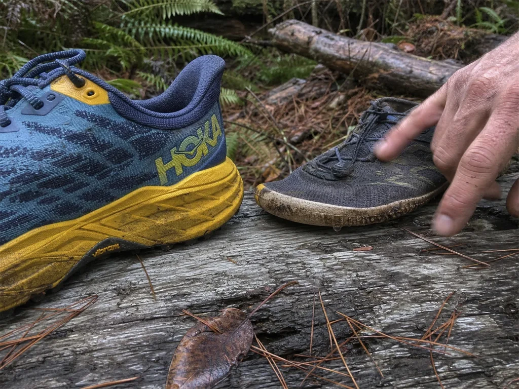 A thick-cushioned trail shoe next to a slim low-stack shoe on a log, showing the ground-feel versus protection trade-off