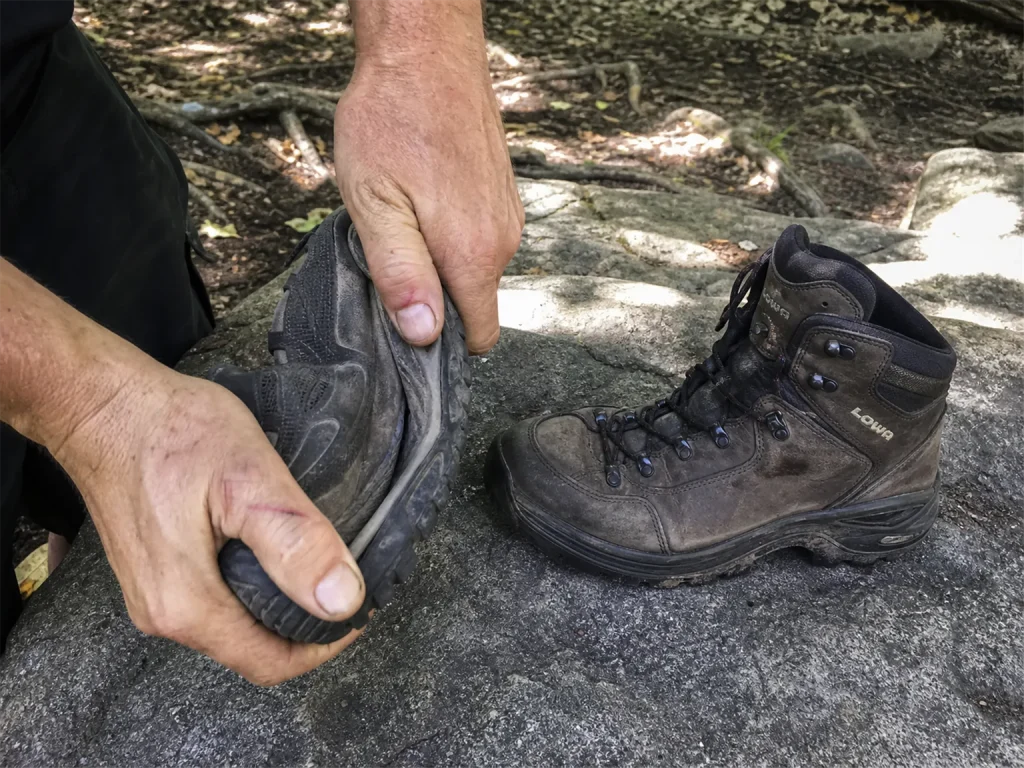 A stiff mid-cut backpacking boot beside a flexible low-cut hiking shoe on a rock, one being twisted to show torsional stiffness