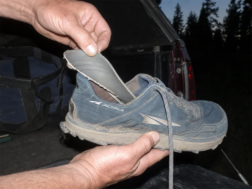 Hands pulling a removable rock plate insert out from under the insole of an Altra Superior shoe at a tailgate at dusk