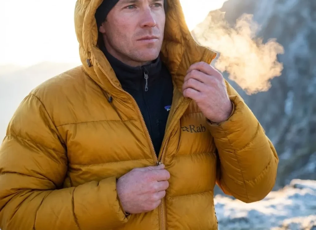 A climber zipping up a thick yellow Rab down jacket, backlit by the first rays of sunrise, showcasing the loft of the insulation and technical fabric.