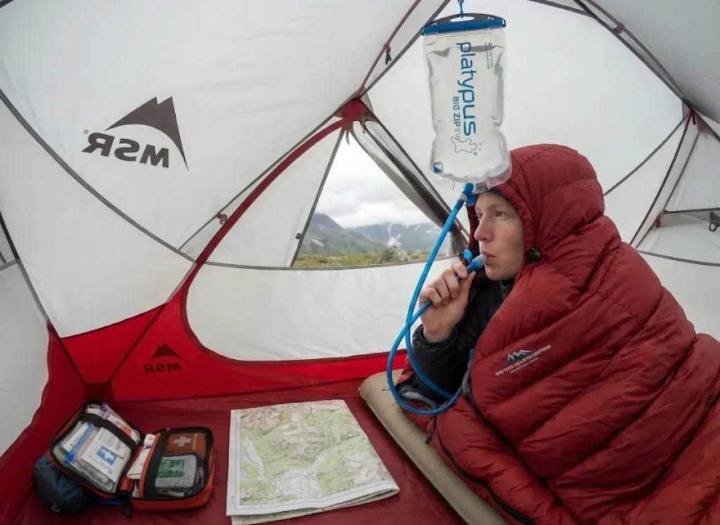 A sick hiker rests inside an MSR tent, sipping electrolytes from a Platypus hydration bladder while resting in a Western Mountaineering sleeping bag.