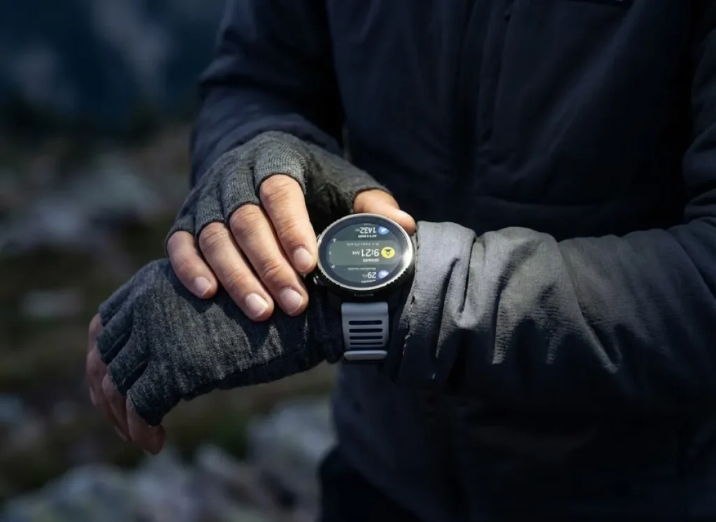 Close-up of a hiker's wrist wearing a Smartwool liner glove and a Suunto GPS watch, illuminated by a headlamp beam in the dark, checking pace statistics.