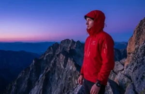 Calculating Start Times: Data-Backed Sunrise Hike Tips A professional hiker in a red Arc'teryx shell stands on a granite ridge during nautical twilight, checking a Garmin watch with a deep blue and violet pre-dawn sky in the background.