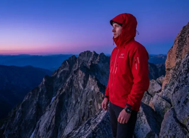 calculating-start-times-sunrise-hike A professional hiker in a red Arc'teryx shell stands on a granite ridge during nautical twilight, checking a Garmin watch with a deep blue and violet pre-dawn sky in the background.