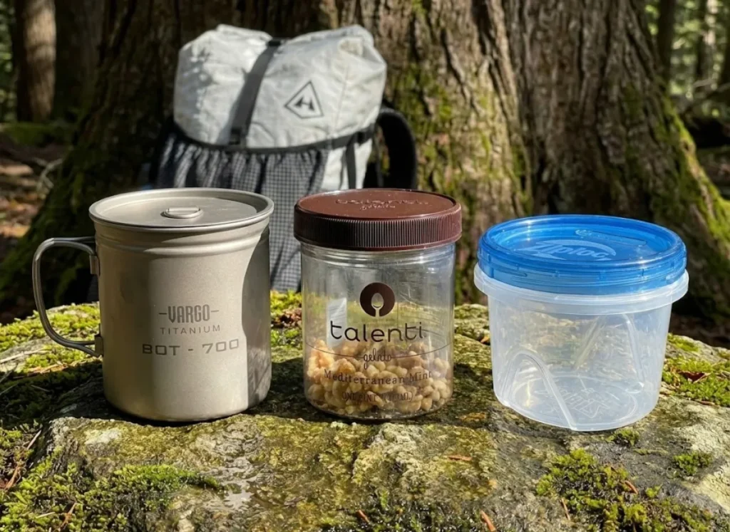 A Vargo titanium pot, Talenti jar, and Ziploc container sitting on a rock next to a Hyperlite Mountain Gear backpack.