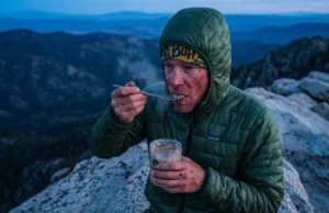Cold Soaking Recipes Ranked by Calories & Clean-Up Hiker on a mountain ridge eating a cold soak meal from a Talenti jar using a Toaks titanium spoon while wearing a Patagonia jacket.