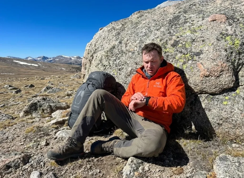 An exhausted hiker wearing an Arc'teryx jacket and checking a Garmin watch rests against a boulder, looking nauseous and fatigued.