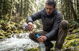 Giardia Symptoms: A Hiker’s Field Guide to Diagnosis A hiker in a Patagonia jacket filters stream water using a Sawyer Squeeze attached to a Smartwater bottle in a dense forest.