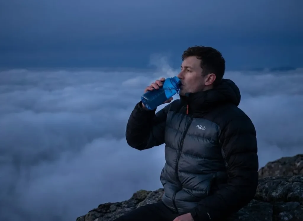A mountaineer in a navy Rab down jacket rests above the clouds during a high-altitude ascent, holding a water bottle with a deep blue sky in the background.