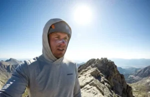 High-Altitude Sun Safety: What Is UPF Clothing Really? Hiker wearing a grey Patagonia Capilene Cool Daily sun hoodie standing on a high altitude granite ridge with intense sunlight overhead.