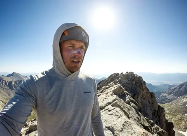 high-altitude-sun-safety-upf Hiker wearing a grey Patagonia Capilene Cool Daily sun hoodie standing on a high altitude granite ridge with intense sunlight overhead.