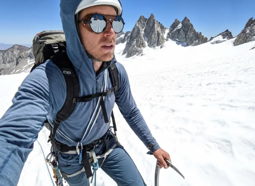 Hiker on a snowfield wearing Outdoor Research sun hoodie and Julbo Vermont Classic glacier glasses reflecting the mountain peaks.