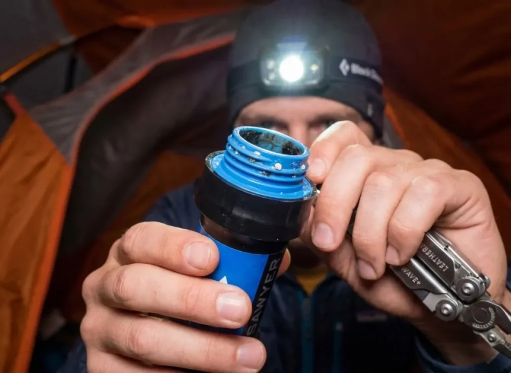 Macro shot of a hiker inspecting the intake threads of a Sawyer Squeeze filter for calcium buildup or biofilm inside a tent.