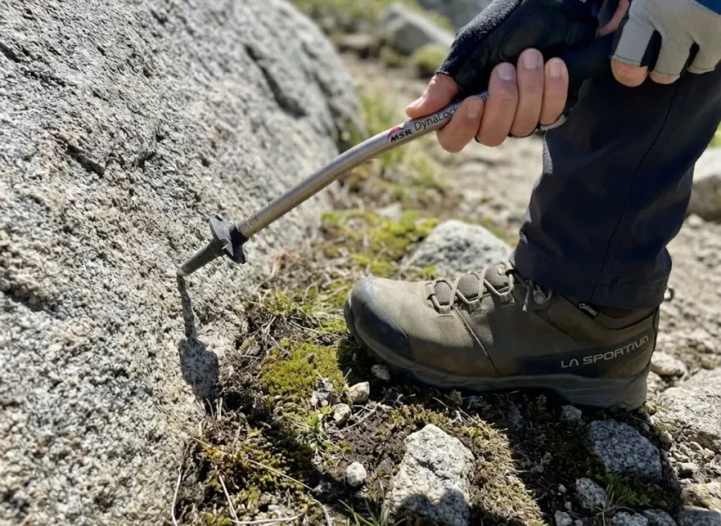 Close up of an MSR DynaLock aluminum trekking pole flexing under pressure against a rock next to a La Sportiva hiking boot.