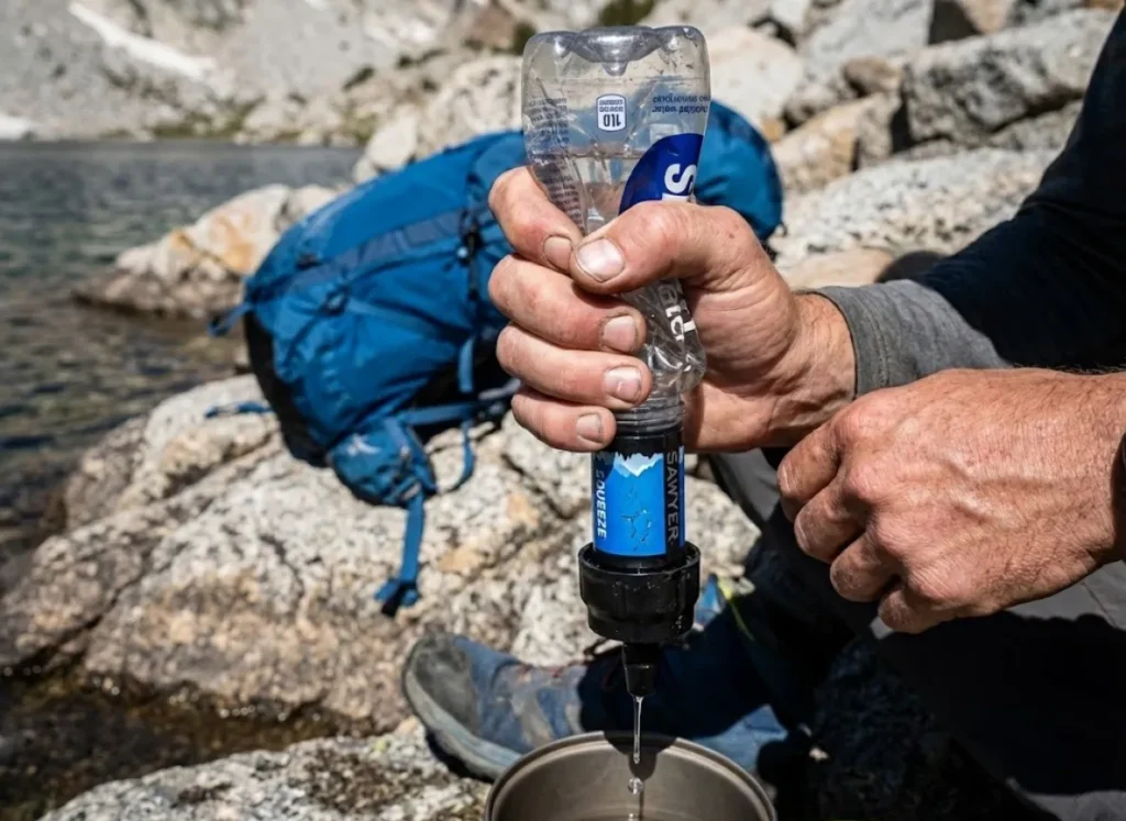 Close up of hiker squeezing a Smartwater bottle with a clogged Sawyer Squeeze water filter attached near an alpine lake.