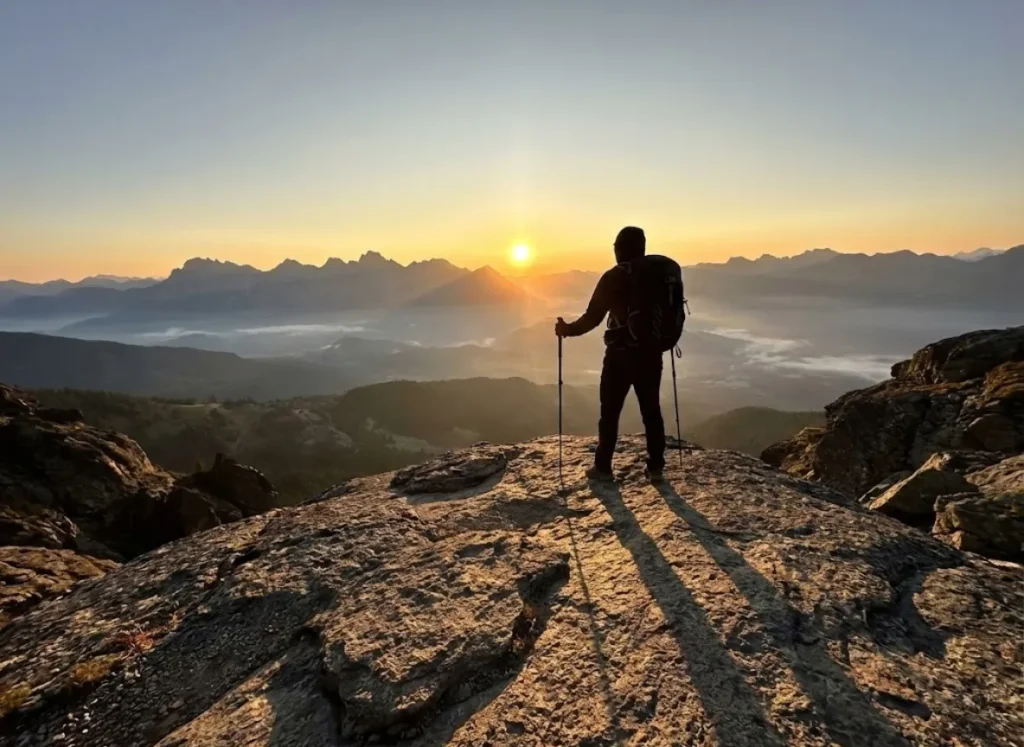 A wide-angle shot of a hiker standing on a mountain peak at the exact moment of sunrise, bathed in golden light with long shadows stretching across the rocky terrain.