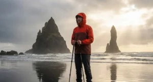 Tide-Safe Guide to the Best Coastal Hikes in the World A hiker in a red Arc'teryx jacket standing on a reflective black sand beach looking out at sea stacks during a dramatic sunset.