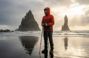 Tide-Safe Guide to the Best Coastal Hikes in the World A hiker in a red Arc'teryx jacket standing on a reflective black sand beach looking out at sea stacks during a dramatic sunset.