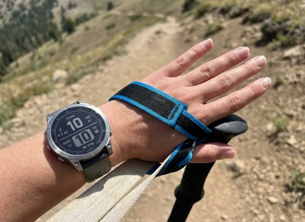 POV shot of a hiker's hand correctly using the wrist strap of a Black Diamond Distance Carbon Z pole with a Garmin Fenix 7 watch visible.