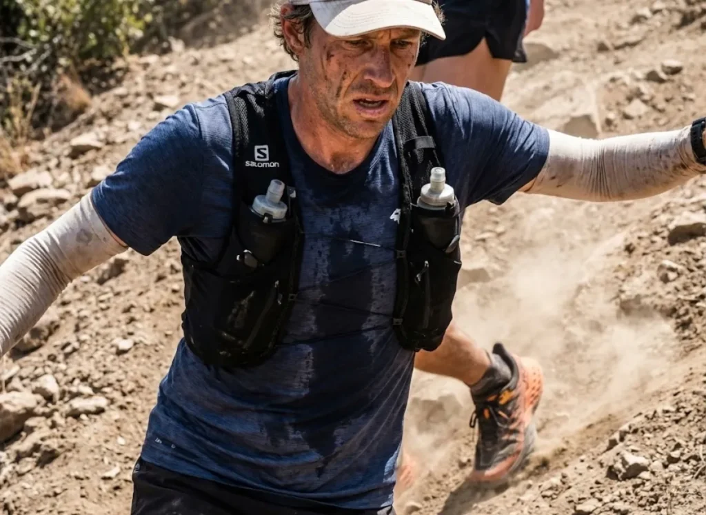 Sweaty trail runner wearing a Salomon hydration vest and a wet Rabbit sun shirt running uphill, showing fabric clinging to skin.
