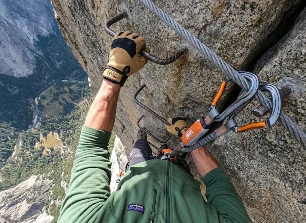 Hiker on a Via Ferrata route using Petzl safety gear and wearing a Patagonia hoodie, looking down a steep drop.