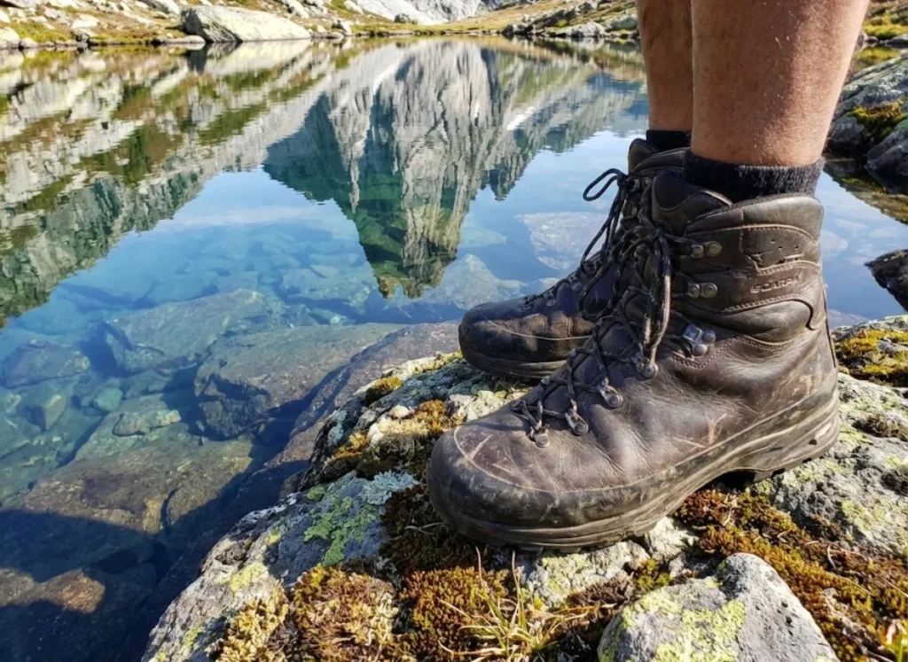 Scuffed Scarpa leather hiking boots standing on granite next to a reflective alpine tarn.