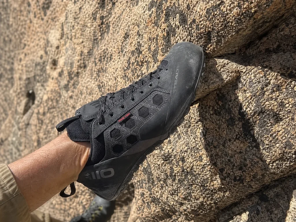 Approach shoe edging onto a small granite foothold on a dry sunlit scramble