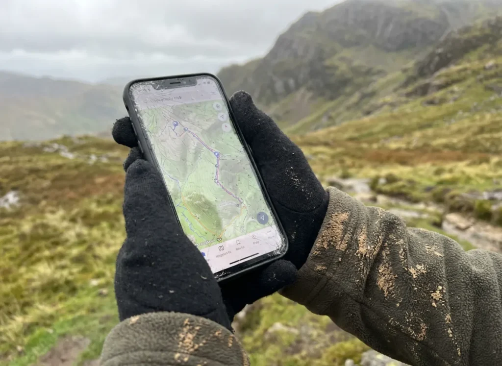 A hiker holding a smartphone showing a GPS map app in a rugged outdoor environment.
