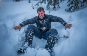 Post Holing Reality Check: Safety, Gear & Etiquette A exhausted winter hiker trapped thigh-deep in snow, struggling to climb out of a posthole in a dense forest.