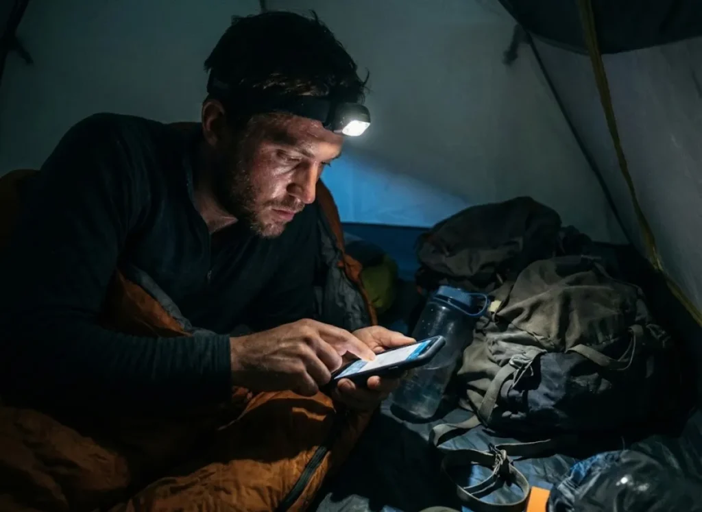 A hiker inside a tent at night, illuminated by a phone screen, writing a thoughtful social media caption after a long day.