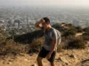Best Urban Hiking Trails in the US: The Field Guide A hiker on a dirt ridge overlooking a smoggy city skyline, wiping sweat from their forehead.