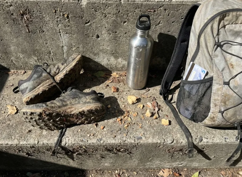 Used trail running shoes and a backpack resting on a concrete wall in a city park.
