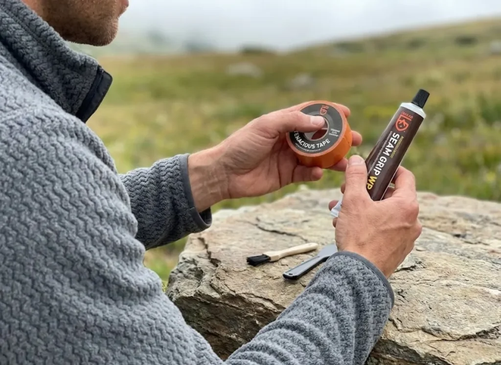 Close-up of a hiker's clean hands comparing Gear Aid Tenacious Tape and Seam Grip adhesive on a rock surface, wearing a Patagonia fleece.