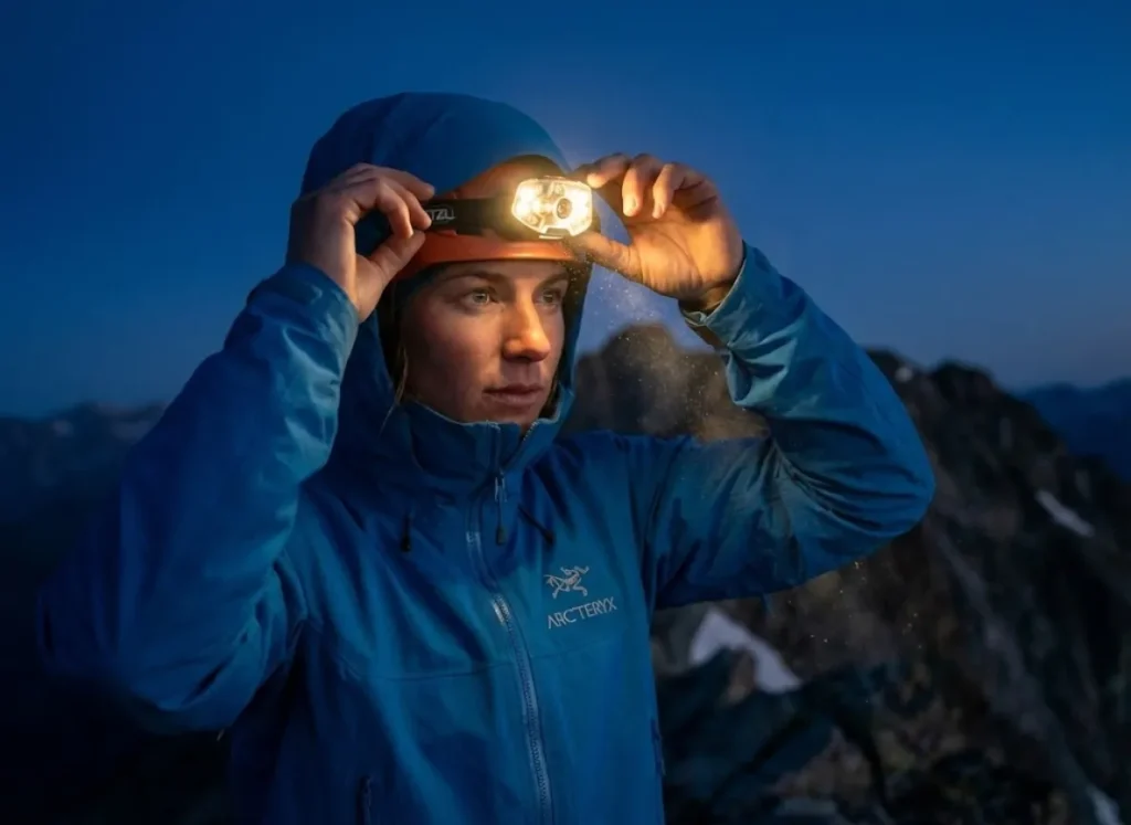 A professional alpinist adjusts a high-tech Petzl headlamp on a dark ridge, the beam cutting through the twilight with visible volumetric light rays.