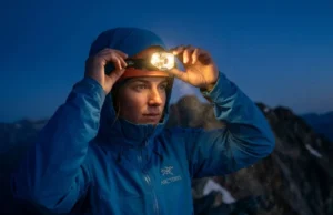 Decoding Lumens vs Candela Headlamp Physics for Hikers A professional alpinist adjusts a high-tech Petzl headlamp on a dark ridge, the beam cutting through the twilight with visible volumetric light rays.