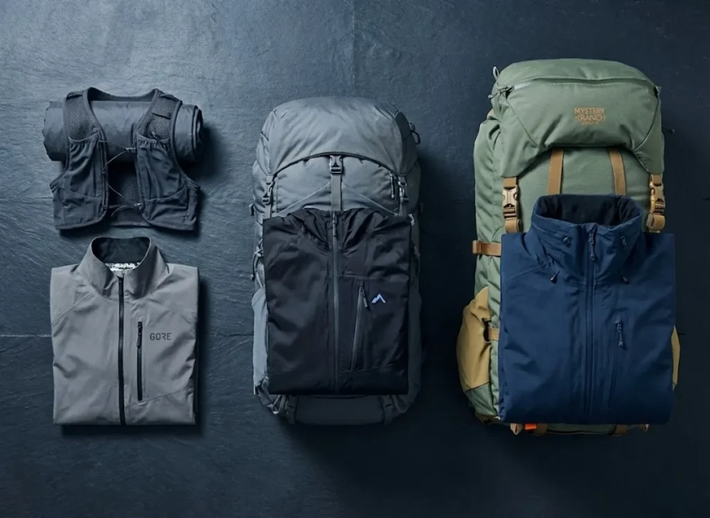 A flat-lay comparison of hiking gear tiers, showing lightweight, midweight, and expedition-weight backpacks paired with their corresponding waterproof jacket types.