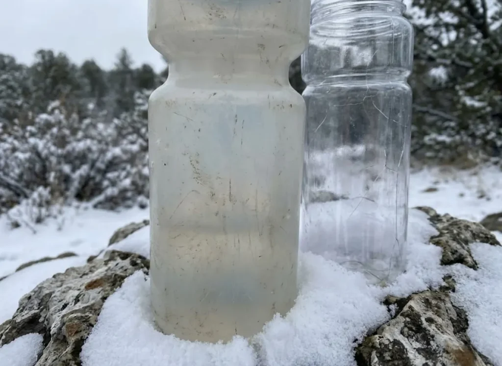 A milky white HDPE water bottle sitting next to a clear plastic bottle on a snow-covered rock.