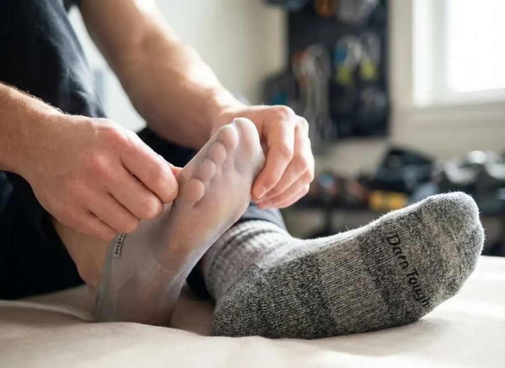 A hiker's clean hands putting on a thin Injinji liner sock, illustrating the double-layer friction prevention system with a thick wool sock ready nearby.