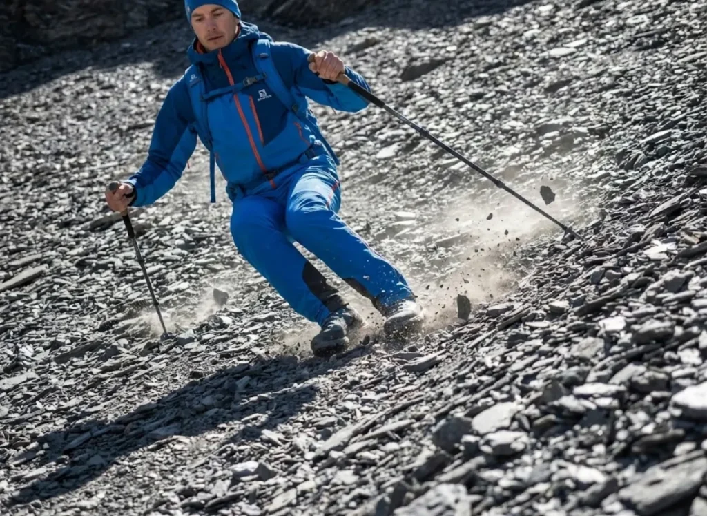 Action shot of a hiker sliding down loose scree using trekking poles for stability, dust frozen in the air, wearing technical Salomon gear.