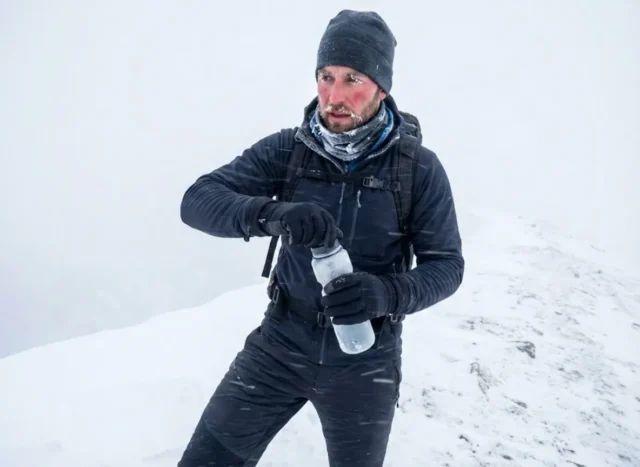 winter-hiking-frozen-water-bottle-struggle A hiker in heavy winter gear struggling to open a frosted water bottle in a snowstorm on a mountain ridge.