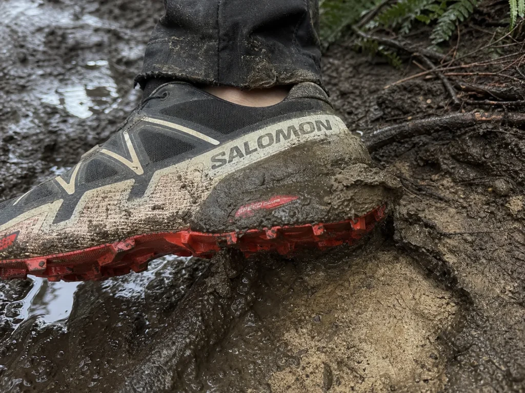 Hiking shoe lug pressing into soft mud, tread biting through the wet surface layer into firmer ground