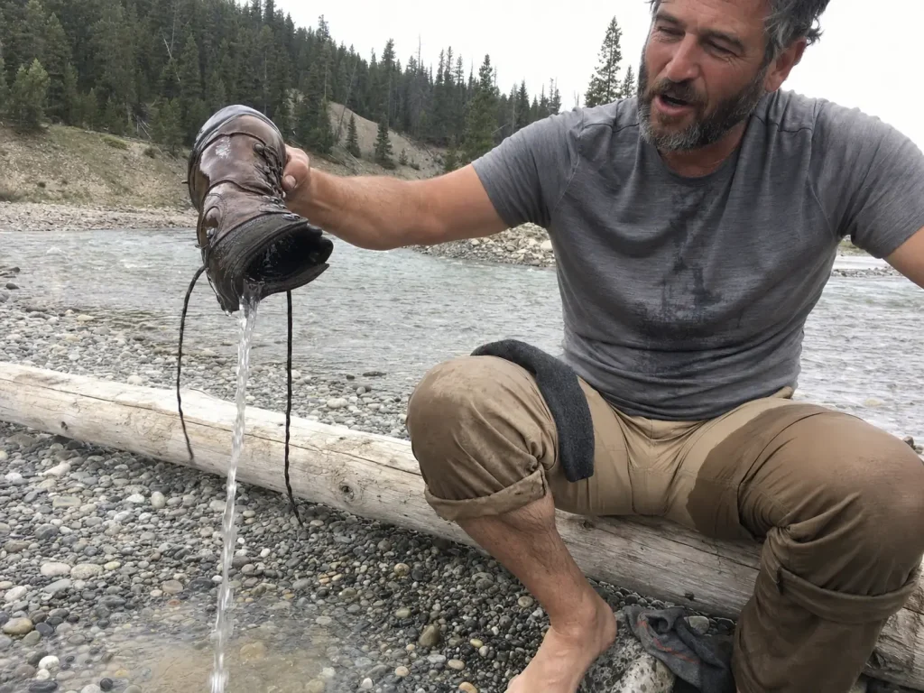 Hiker sitting on a gravel bank tipping water out of a waterproof boot after a stream crossing