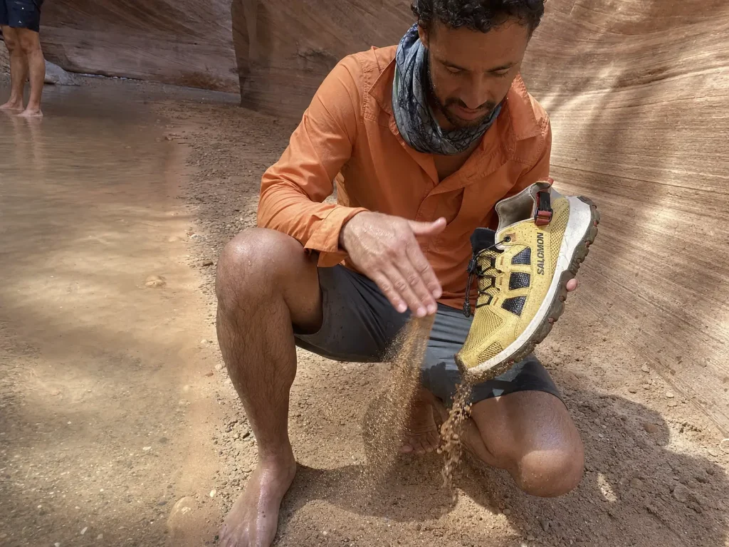 Hiker shaking sand out of an anti-debris mesh drainage shoe in a sandy desert canyon creek